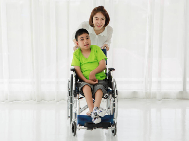Jovem positivo asiático feminino cuidador rolando cadeira de rodas com adorável sorrindo menino étnico que vive com deficiência de paralisia cerebral na clínica moderna - Foto, Imagem