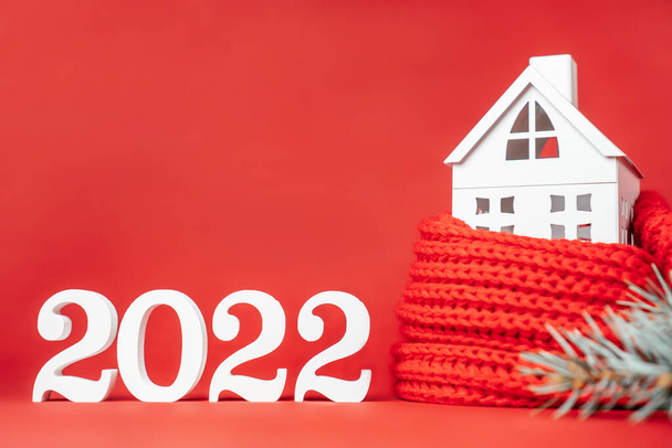 Šťastný Nový rok2022. Bílá dřevěná čísla 2022 stojan s bílou hračkou dům zabalený v červené šály a vánoční strom větev na červeném pozadí. Veselé Vánoce. Kopírovat prostor - Fotografie, Obrázek