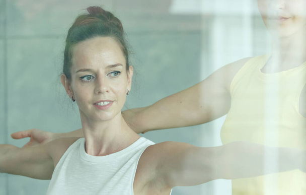 Weibliche Yoga-Meisterin bringt der Schülerin bei, grundlegende Übungen zu beginnen und hilft ihr, die richtige Körpergeste zu arrangieren. Von Außenspiegelwand mit Reflexion aufgenommen. - Foto, Bild
