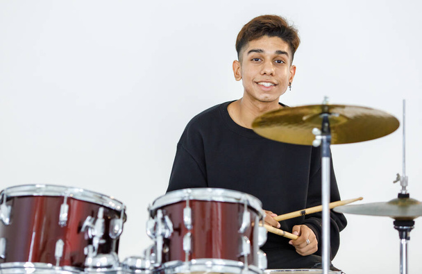 Портретний знімок підліткового барабанщика, який грає музику. Молодий музикант грає на барабані, дивлячись на камеру і посміхаючись. Молодший студент грає на інструменті з білим тлом
 - Фото, зображення