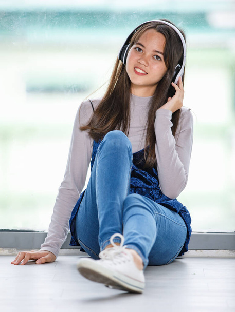 Portré felvétel fiatal thai-török tinédzserről, aki fejhallgatóval zenét hallgat. Junior vegyes fajú lány élvezi a zenét kézzel érintse meg a fejhallgató és csukott szemmel gesztus, miközben ül a padlón - Fotó, kép