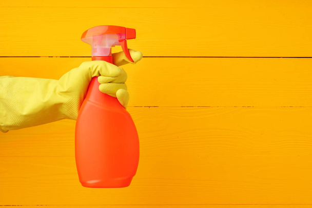 Hand in glove holding household detergent spray against yellow wooden background - Fotó, kép