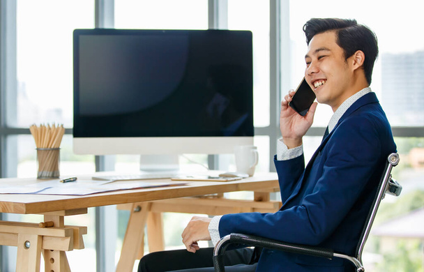 アジアの若いビジネスマンは、デスクに座って、スマートフォンを使用してデスクトップコンピュータの近くで通話や通信を行う. - 写真・画像