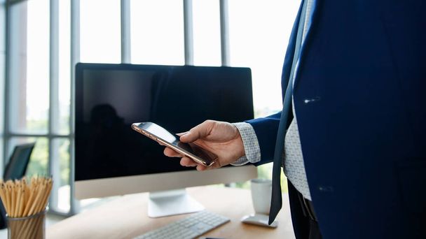 Nierozpoznawalne zdjęcie biznesmena w niebieskim garniturze trzymającego i używającego smartfona w biurze z rozmytym komputerem stacjonarnym w tle. - Zdjęcie, obraz