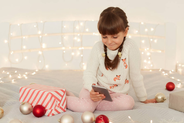 Cute little girl schoolgirl wearing casual attire sitting on bed at home holding mobile phone in hands, checking social networks with children content. - Φωτογραφία, εικόνα