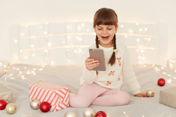 Portrait of happy little adorable girl wearing white casual sweater sitting on bed with cell phone in hands, having video call with friends, having excite expression, celebrating Christmas and new year - Photo, image