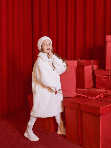 Cute little girl in white coat and had by the huge red christmas presents boxes on red background. Holidays, shopping, fun, fashion - Фото, зображення