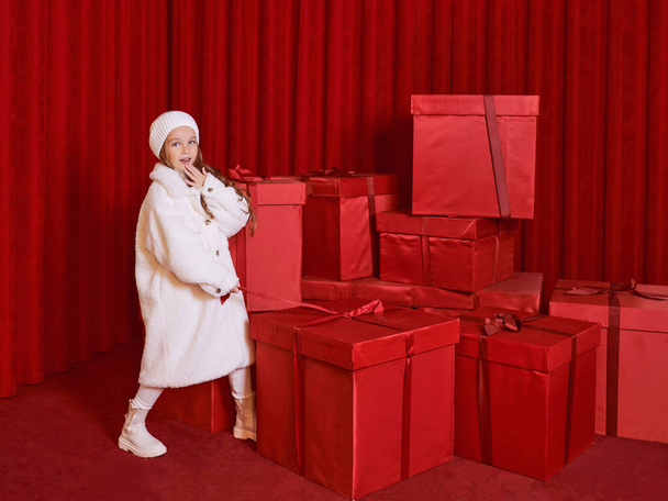 Cute little girl in white coat and had by the huge red christmas presents boxes on red background. Holidays, shopping, fun, fashion - Photo, image