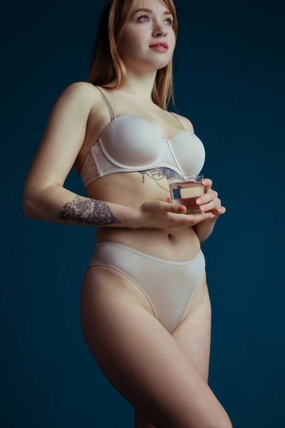 Portrait of young woman in nude color underwear with glass of water isolated on dark blue studio background. Natural beauty concept. - Fotoğraf, Görsel