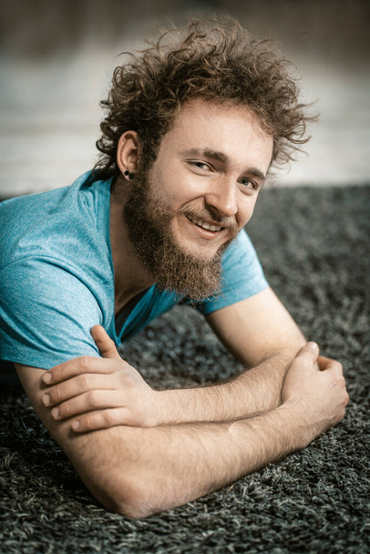 Attractive Caucasian Guy in a Blue T-shirt Relaxed on the Carpet. Smiling Man Resting on the Carpet After Funny Party. Close-up  - Фото, зображення