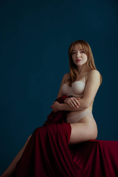 Classic portrait of young beautiful red-headed woman in nude color underwear posing isolated over blue background. - Foto, imagen