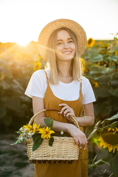 Eine Frau steht mit einem Blumenkorb in der Hand durch ein Sonnenblumenfeld. Sie lächelt und blickt in die Kamera. Sonnenuntergang im Hintergrund. - Foto, Bild
