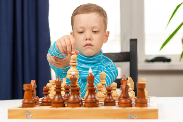 Aranyos hétéves kisfiú, aki otthon sakkozik egy fehér fa asztalon. Szelektív fókusz. Közelkép. Portré - Fotó, kép