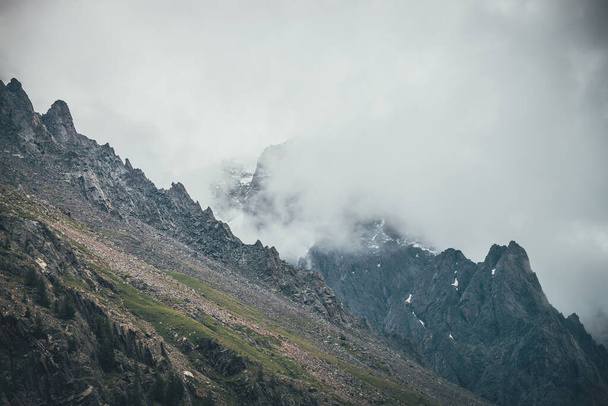 灰色の曇りの空に低い雲の中に暗い岩の山の頂上と暗い大気のシュールな風景。高い頂点に灰色の低い雲。低い雲の中に雪と高い黒い岩。シュールレアリズムの暗い山. - 写真・画像