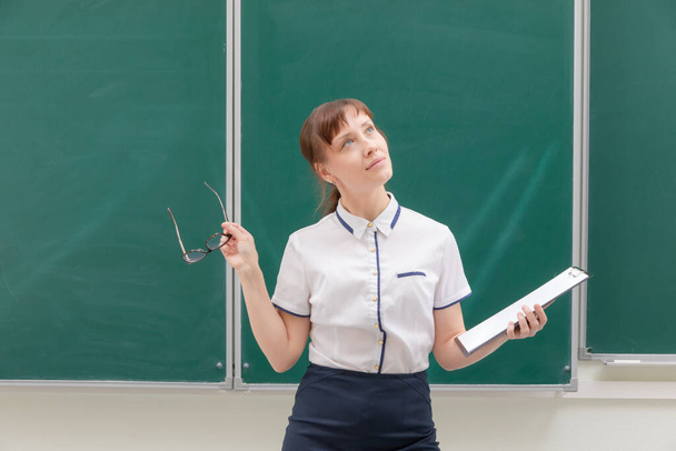 schooljuf jonge mooie vrouw in een witte blouse met een map en bril in haar handen in de klas op de achtergrond van het schoolbord. portret - Foto, afbeelding