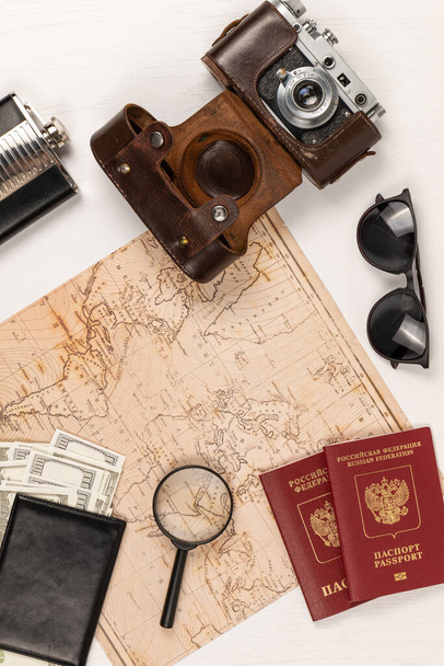 Dünya haritası, iki pasaport, siyah deri bir cüzdanda para deri bir çantada eski bir kamera ve beyaz tahta bir masada güneş gözlüğü. Yukarıdan bak. Seçici odaklanma. Yazı - pasaport Rusya Federasyonu - Fotoğraf, Görsel