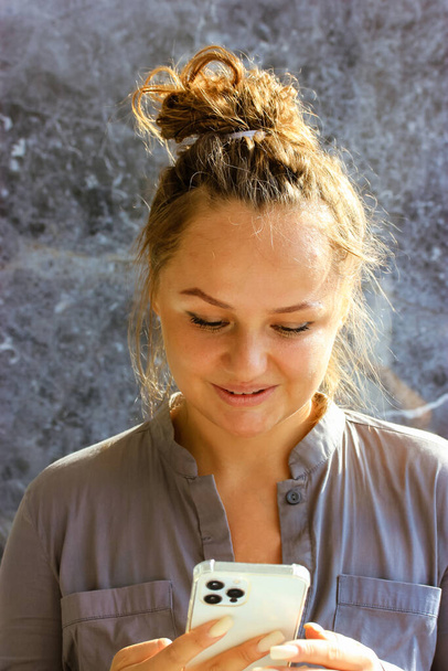 A young, pretty, smiling woman holding a cell phone is typing a text message, chatting, shopping online. Vertical portrait of a girl, lady with her hair in a bun in sunlight. Pleasant communication. - Фото, зображення