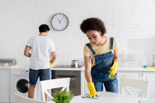 african american woman cleaning dining table with rag near blurred boyfriend  - Fotografie, Obrázek