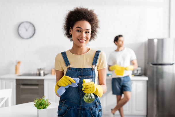 cheerful african american woman holding spray bottle and rag near blurred man in kitchen  - Photo, Image
