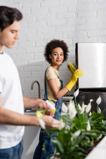 donna afro-americana sorridente che tiene la spugna vicino al frigorifero vicino all'uomo offuscato che innaffia le piante in cucina  - Foto, immagini