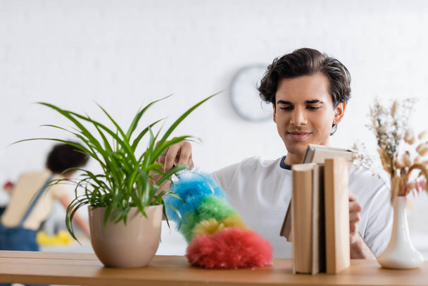 young man cleaning wooden shelf with dust brush near plants and books - Foto, Bild