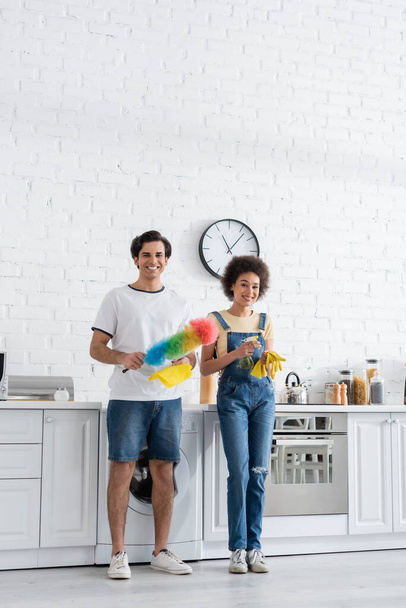 full length of cheerful young man holding dust brush near african american girlfriend with rubber gloves - Foto, afbeelding