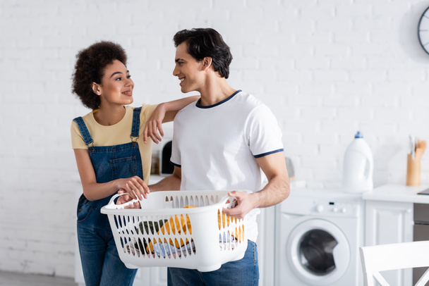 happy interracial couple looking at each other near basket with dirty laundry  - Photo, Image