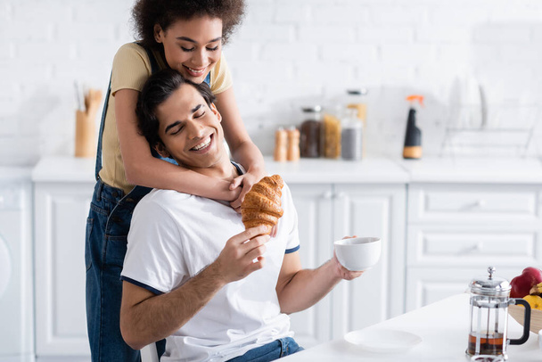happy african american woman hugging boyfriend with cup of tea and croissant  - Foto, Bild