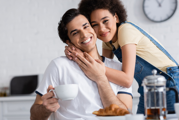 joyful african american woman hugging boyfriend with cup of tea in kitchen - Valokuva, kuva
