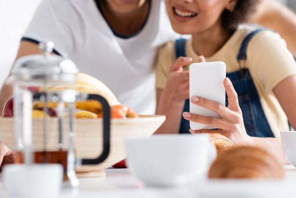 cropped view of happy interracial couple using smartphone during breakfast  - Fotografie, Obrázek
