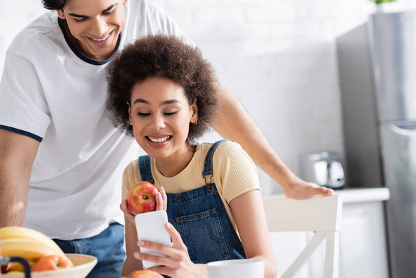 cheerful interracial couple looking at smartphone during breakfast  - Фото, зображення