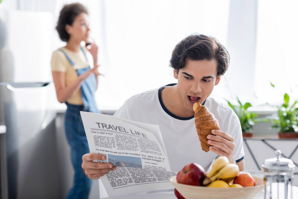 hombre joven comiendo croissant y leyendo el periódico de la vida de viaje cerca borrosa mujer afroamericana  - Foto, imagen