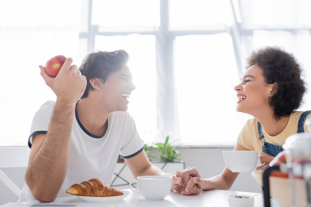 вид сбоку счастливой многонациональной пары, держащейся за руки во время завтрака - Фото, изображение