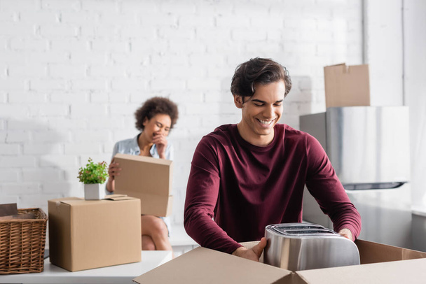 cheerful young man unpacking box with toaster near blurred african american girlfriend  - Photo, Image