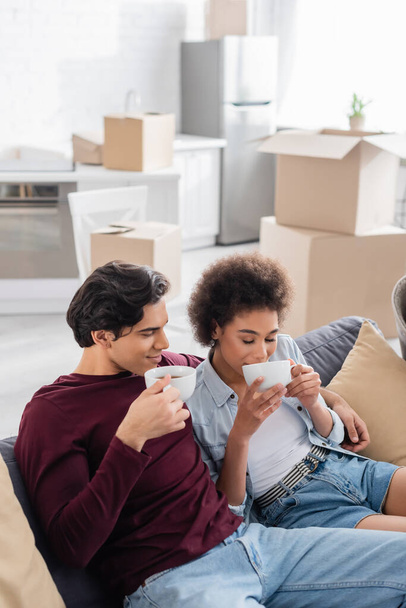 happy multiethnic couple drinking tea while resting on couch during relocation  - Foto, imagen