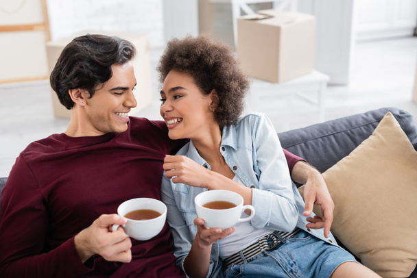 fröhliches multiethnisches Paar mit Tassen Tee, während es sich auf der Couch ausruht  - Foto, Bild