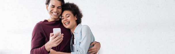 happy man using smartphone and hugging smiling african american girlfriend, banner - Foto, Imagem