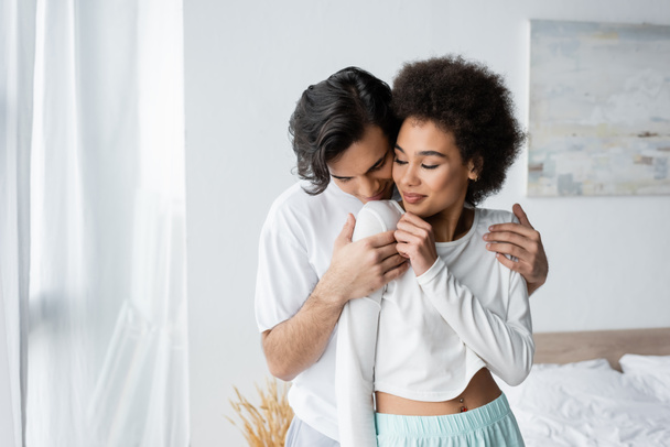 man hugging smiling african american girlfriend in bedroom  - 写真・画像