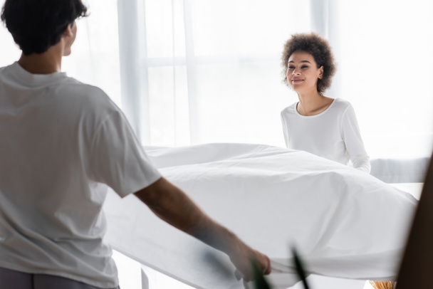 positive african american woman arranging bed sheet with boyfriend  - Fotografie, Obrázek