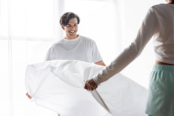 happy man arranging bed sheet with african american girlfriend  - 写真・画像