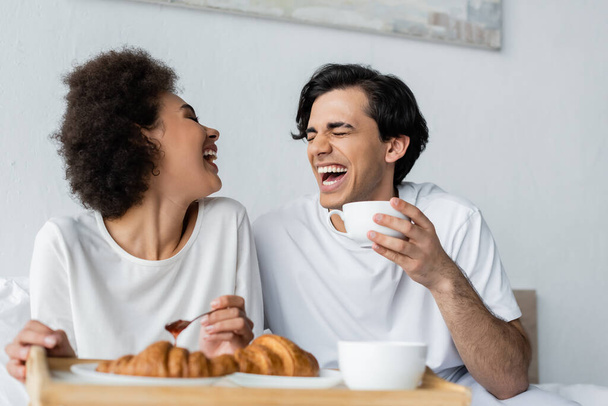 fröhliche afrikanisch-amerikanische Frau hält Löffel mit Marmelade neben Freund mit Tasse beim Frühstück im Bett  - Foto, Bild