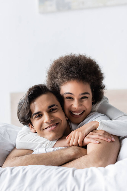 alegre interracial pareja sonriendo mientras abrazo en cama - Foto, Imagen