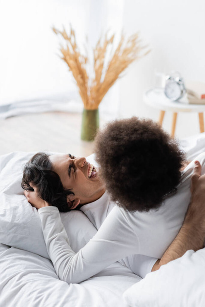 curly african american woman stroking hair of cheerful man on bed - Photo, image