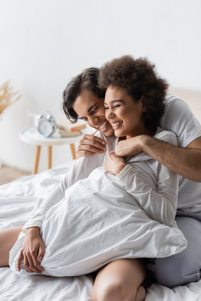 cheerful man hugging pleased african american woman on bed - 写真・画像