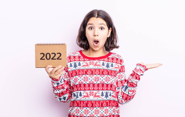 pretty young woman looking surprised and shocked, with jaw dropped holding an object. 2022 calendar concept - Фото, изображение