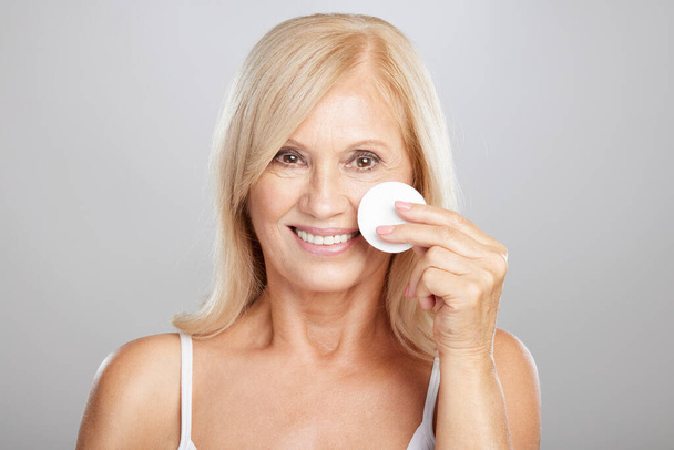 A blond senior woman is removing makeup with a cotton pad. - Fotó, kép