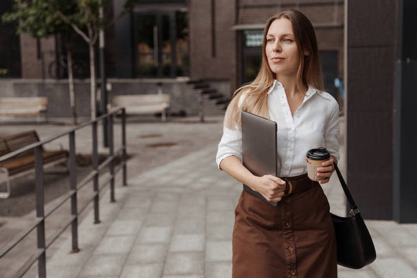  Junge Geschäftsfrau mit Kaffee und Laptop im Hintergrund des modernen Bürogebäudes  - Foto, Bild