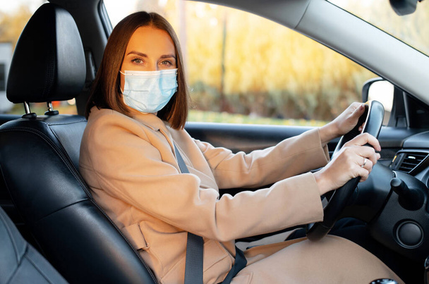 Young woman in autumn coat and in protective medical mask driving a car - Fotó, kép