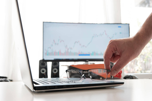 Clear reading glasses, black rim, on a laptop keypad. Blurred large computer monitor in the background showing stock market graph. Male hand grabbing the glasses. - Photo, Image