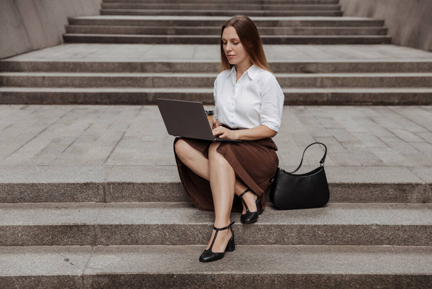 Joven mujer de negocios sentada en una escalera con un ordenador portátil del moderno edificio de oficinas de fondo  - Foto, Imagen